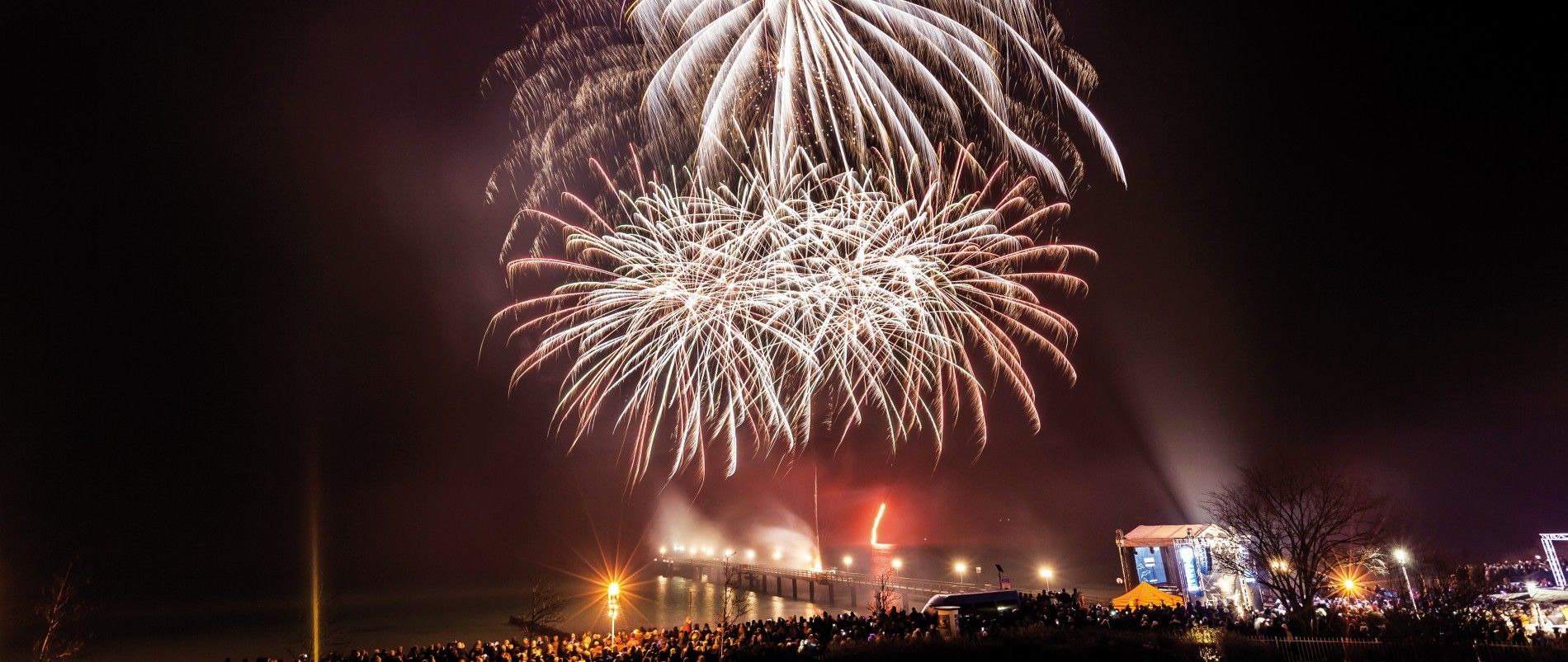 Großes Feuerwerk über einer Menschenmenge bei Nacht, mit einer beleuchteten Bühne in der Nähe.