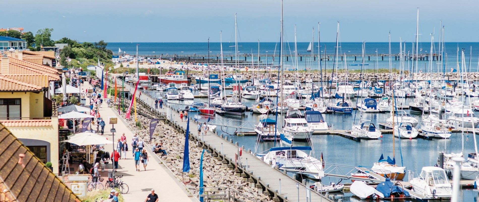 Hafen mit vielen Segelbooten und einer belebten Promenade bei sonnigem Wetter.
