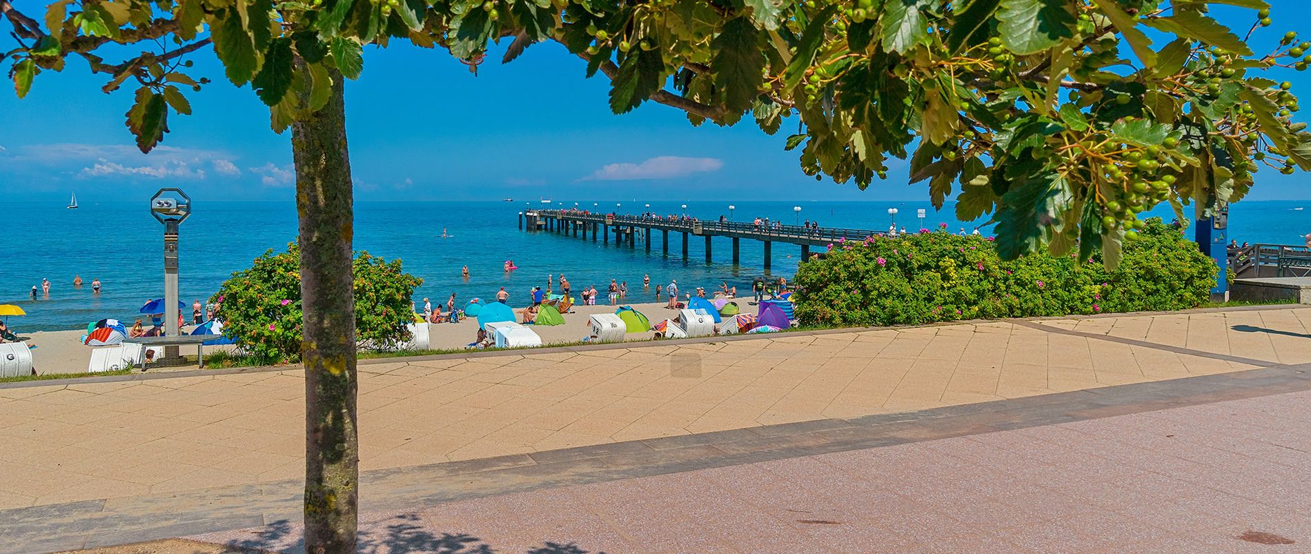 Strand mit bunten Strandkörben, einem Pier und Menschen im Wasser an einem sonnigen Tag.