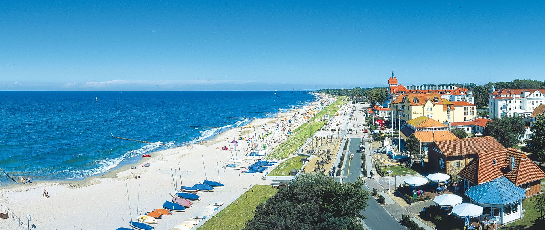 Panoramablick auf einen Strand an der Ostsee mit bunten Sonnenschirmen und Segelbooten, daneben eine Promenade mit historischen Gebäuden.