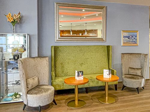 Gemütliche Sitzecke mit grünem Sofa, zwei grauen Sesseln und runden Holztischen in einem modern eingerichteten Raum mit großen Spiegel und Gemälden an der Wand.