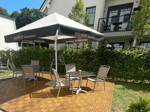 Ein Außenbereich mit einem Mövenpick-Sonnenschirm, darunter stehen Tische und Stühle auf einem Holzboden. Im Hintergrund ist eine Hecke und ein Gebäude zu sehen.