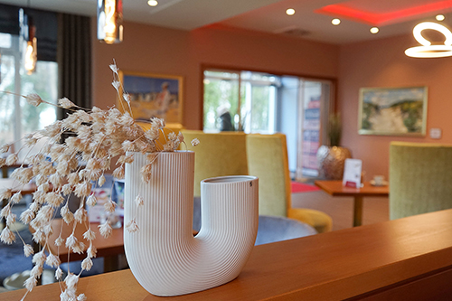 Eine moderne, weiße Vase in ungewöhnlicher Form steht auf einer Theke mit getrockneten Blumen. Im Hintergrund ist ein warm gestalteter Raum mit Tischen, Stühlen und Gemälden an den Wänden.