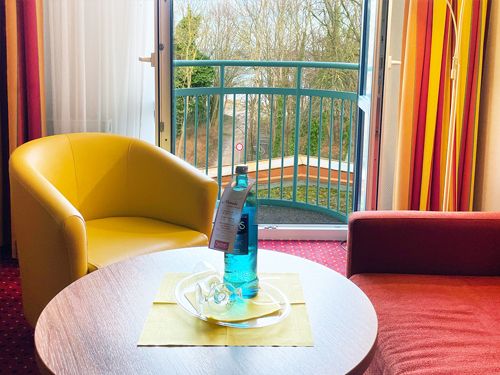 Hotelzimmer mit gelbem Sessel und rotem Sofa, Blick auf Balkon mit Bäumen.