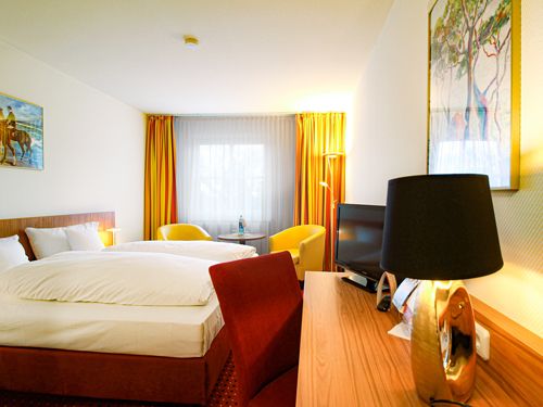 Gemütliches Hotelzimmer mit Doppelbett, rotem Stuhl, gelben Sesseln, Schreibtischlampe und Fernseher.