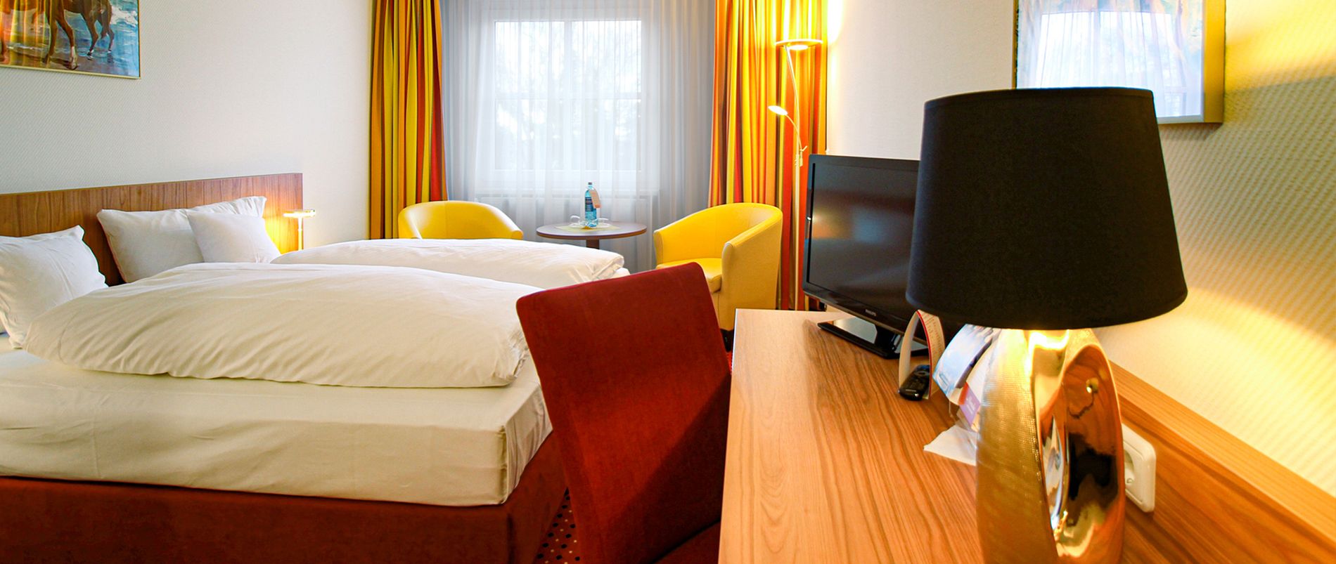 Gemütliches Hotelzimmer mit Doppelbett, gelben Sesseln und Schreibtischlampe.